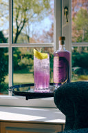 The Illusionist Rosemary Fizz ready-to-drink cocktail garnished with rosemary and lemon, served on a black tray beside the bottle by a sunlit window.
